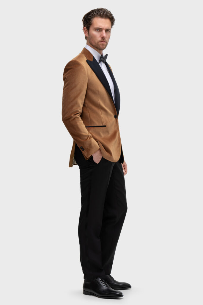 Ein Mann in hellbraunem Samtblazer, schwarzer Hose und Fliege steht mit einer Hand in der Tasche da - italienischer Stil und die Eleganz von Hamburgs feinsten Maßanzügen.