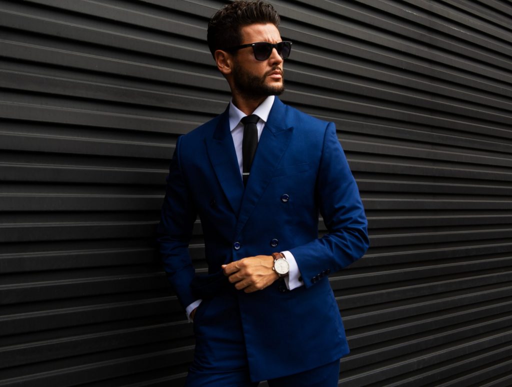 Ein Mann im blauen Konfektions-Anzug und mit Sonnenbrille steht selbstbewusst vor einer schwarz strukturierten Wand und verkörpert moderne Hamburger Herrenmode und die Stilsicherheit eines erfahrenen Herrenausstatters.