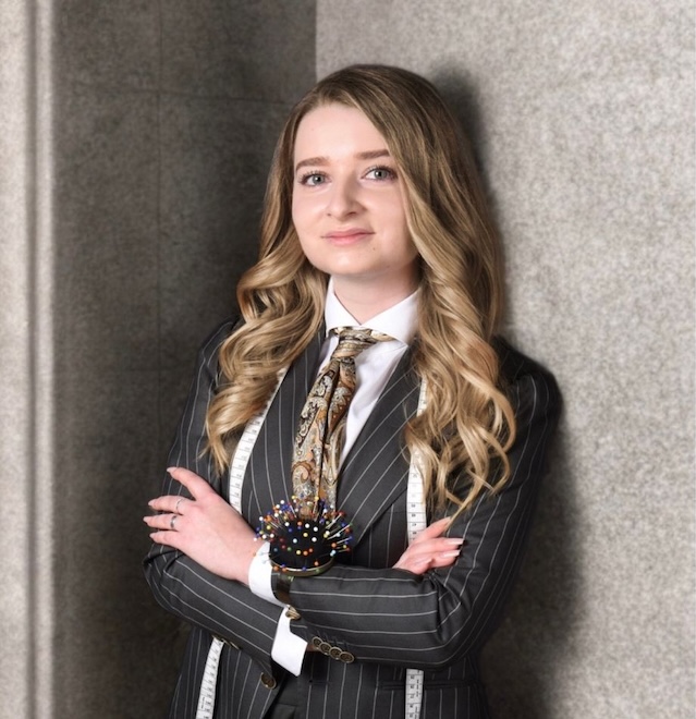 Frau, Lara Lukomski, im Nadelstreifenanzug mit langen blonden Haaren, die Arme verschränkt, steht vor einer grauen Wand - eine elegante Anspielung auf Hamburgs feinste Herrenmode und Herrenausstatter-Stil.