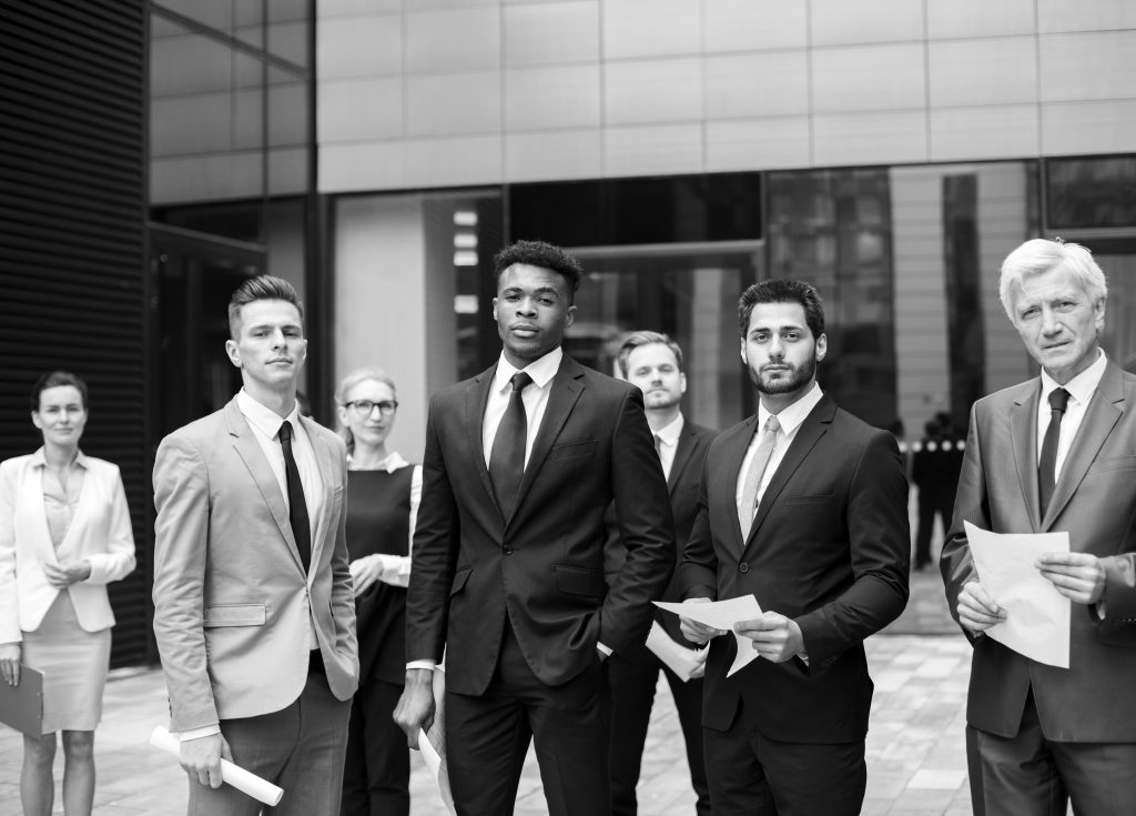 Eine Gruppe von Geschäftsleuten in Maßanzügen steht im Freien zusammen, hält Papiere in der Hand und schaut selbstbewusst in die Kamera.