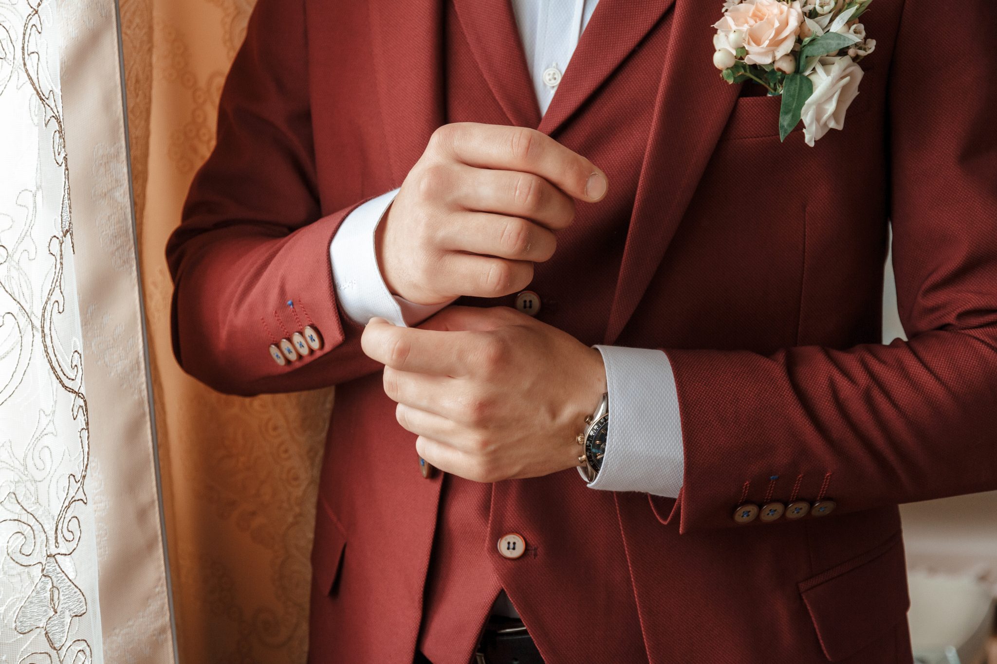 Mann im italienischen burgunderroten Hochzeitsanzug, der seine Manschette zurechtrückt, mit einer Boutonniere am Revers, steht neben einem Vorhang.