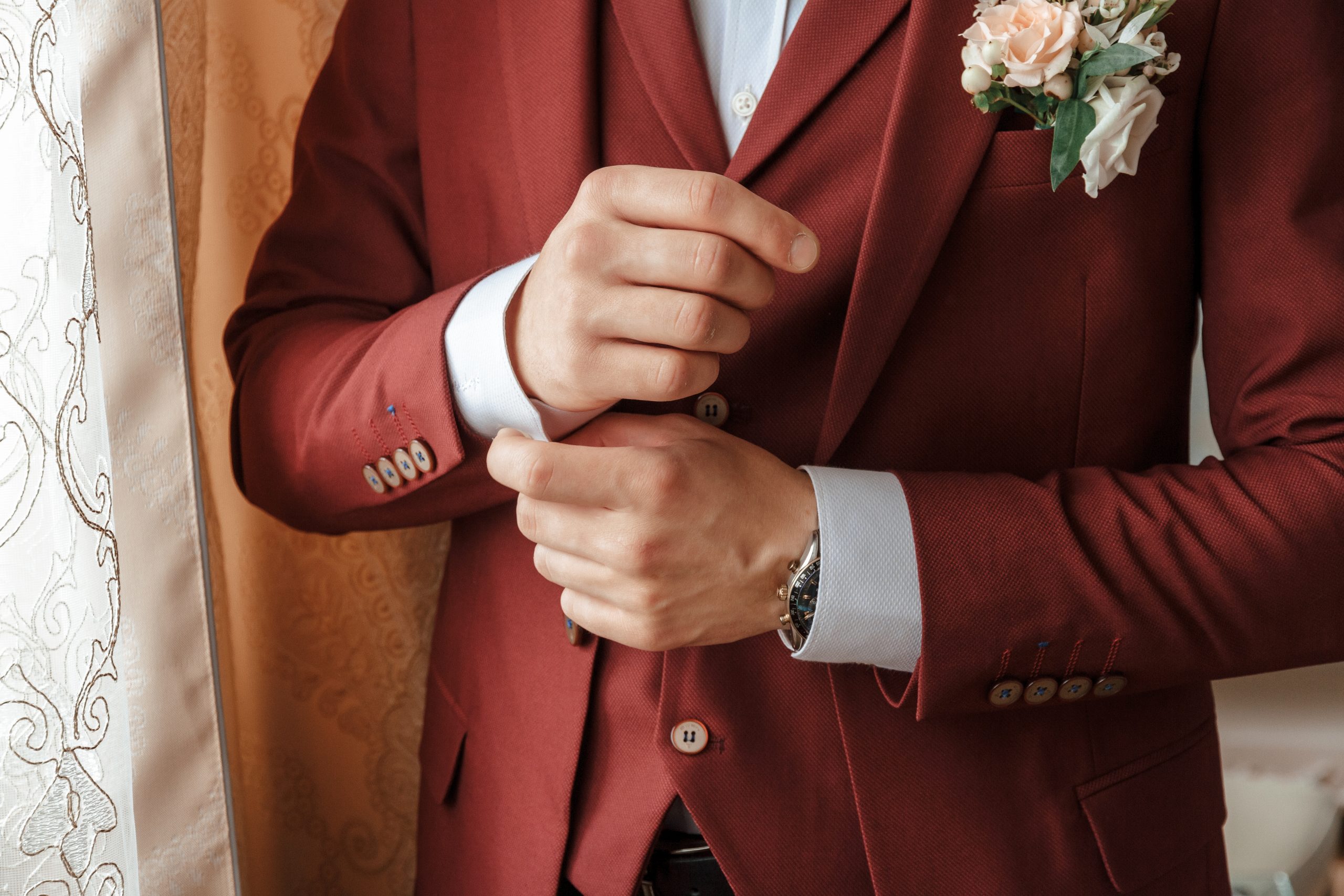 Mann im italienischen burgunderroten Hochzeitsanzug, der seine Manschette zurechtrückt, mit einer Boutonniere am Revers, steht neben einem Vorhang.