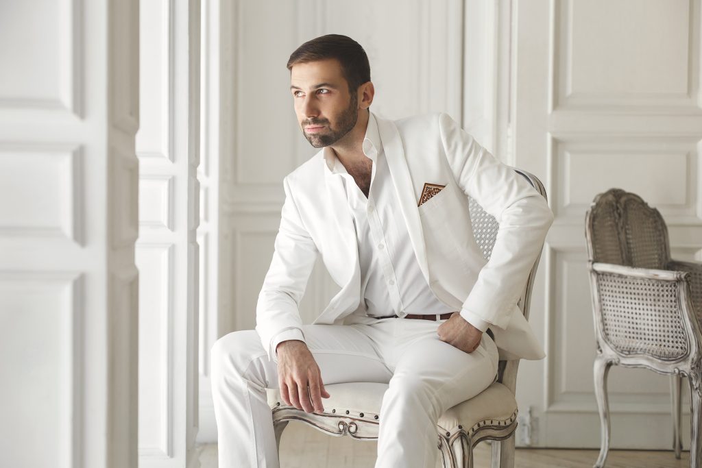 Mann im eleganten, italienischen Hochzeitsanzug sitzt nachdenklich auf einem Stuhl in einem hellen, eleganten Raum und schaut zur Seite.