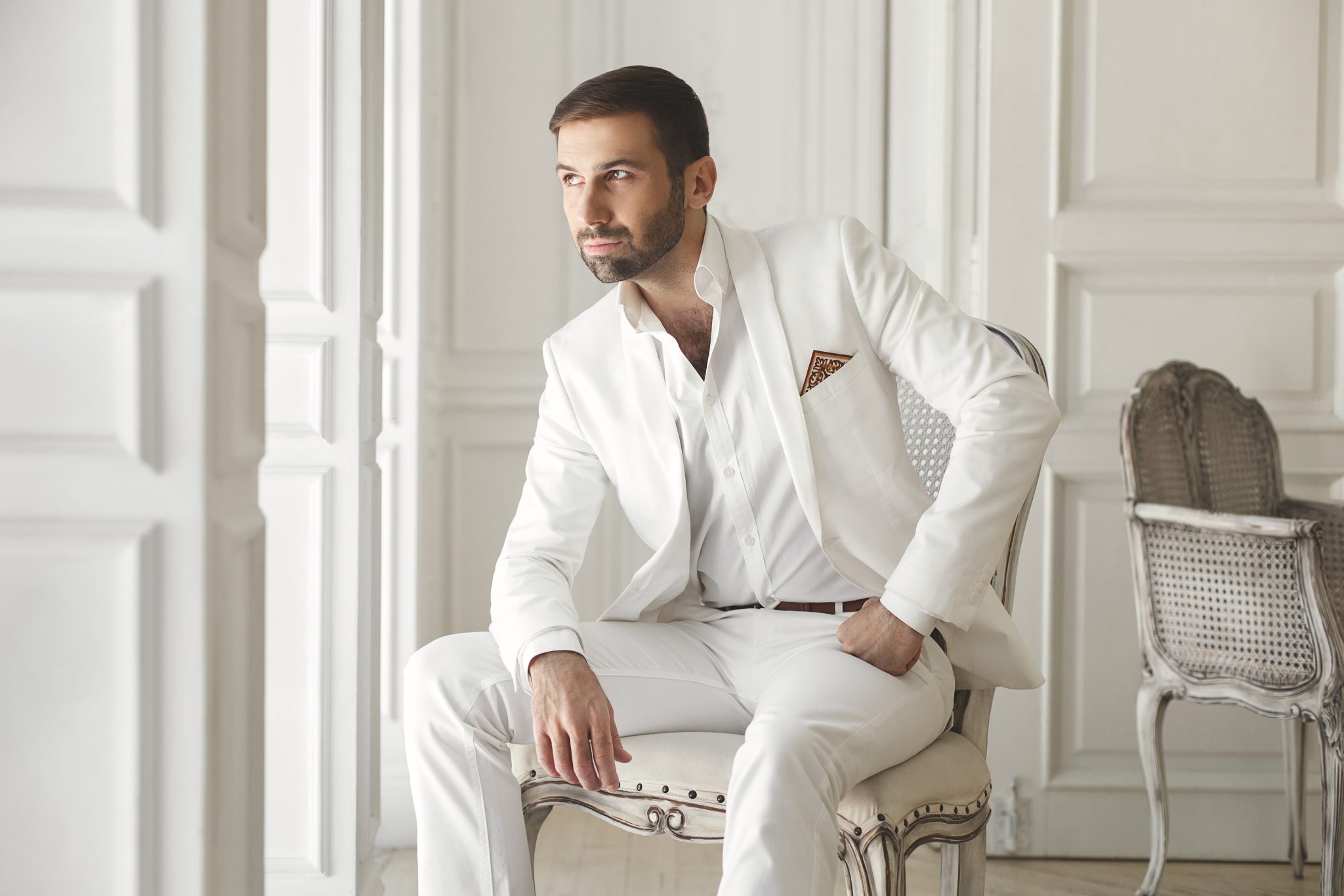 Mann im eleganten, italienischen Hochzeitsanzug sitzt nachdenklich auf einem Stuhl in einem hellen, eleganten Raum und schaut zur Seite.