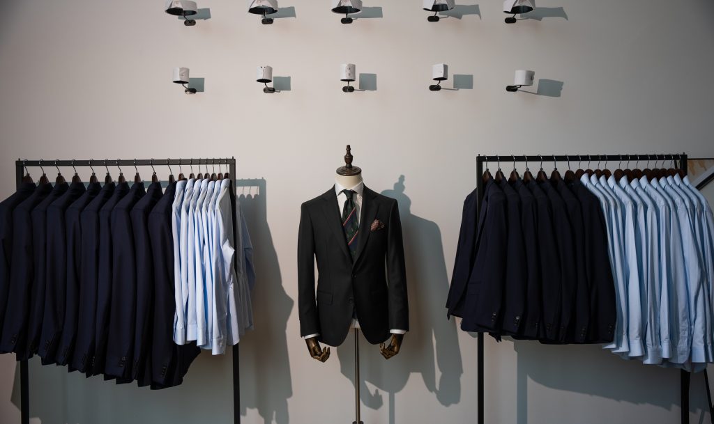 In einem stilvollen Bekleidungsgeschäft steht eine Varazio-Schaufensterpuppe im Anzug zwischen Kleiderständern mit Anzügen und Hemden, beleuchtet von modernen Wandleuchten über ihr.