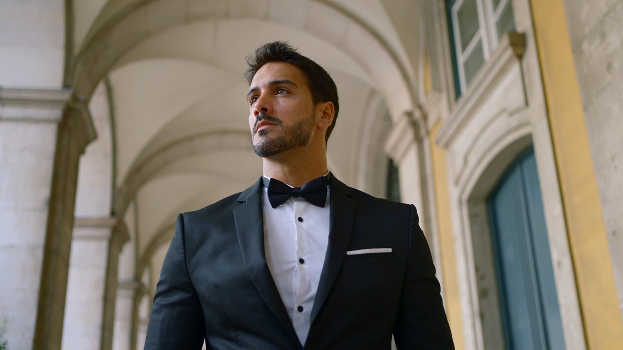 Ein Mann in einem schwarzen Anzug steht selbstbewusst unter einem gewölbten Gang, den Blick zur Seite gerichtet – bereit für die bevorstehende Hochzeit.