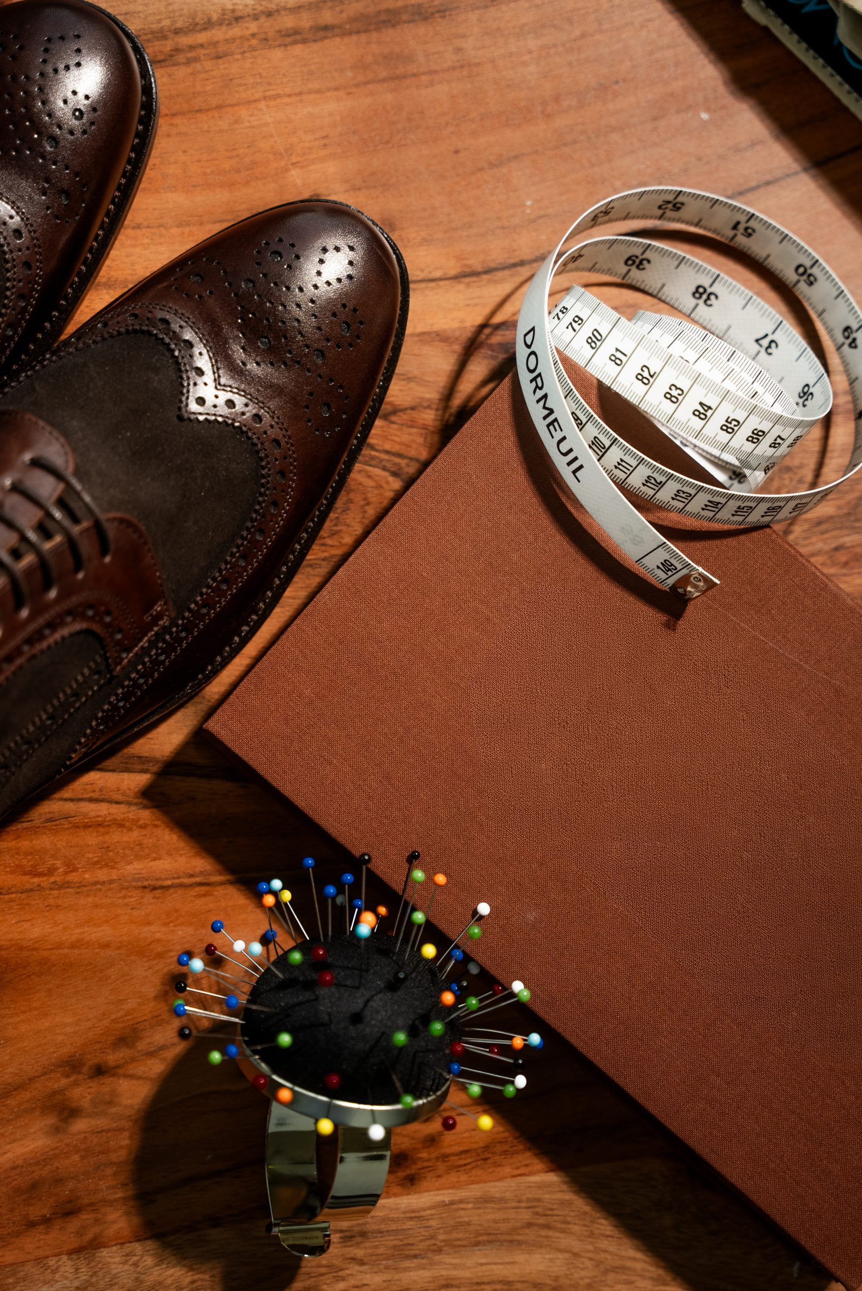 Braune Kleiderschuhe, ein geschlossenes braunes Buch, ein Nadelkissen mit Stecknadeln und ein Maßband auf einer Holzfläche.